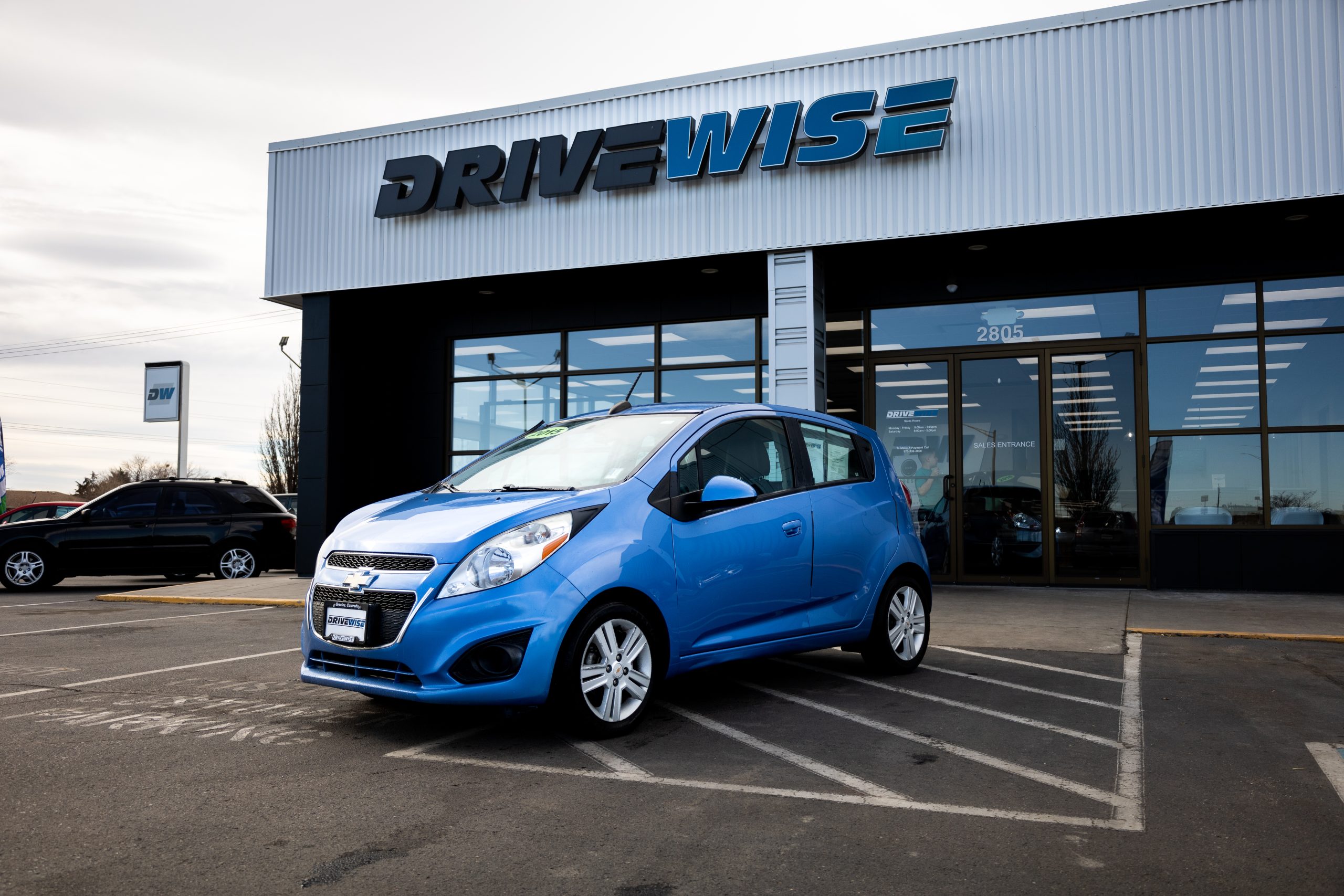 A blue Chevrolet Spark hatchback parked in front of the DriveWise Budget Cars building in Greeley, Colorado